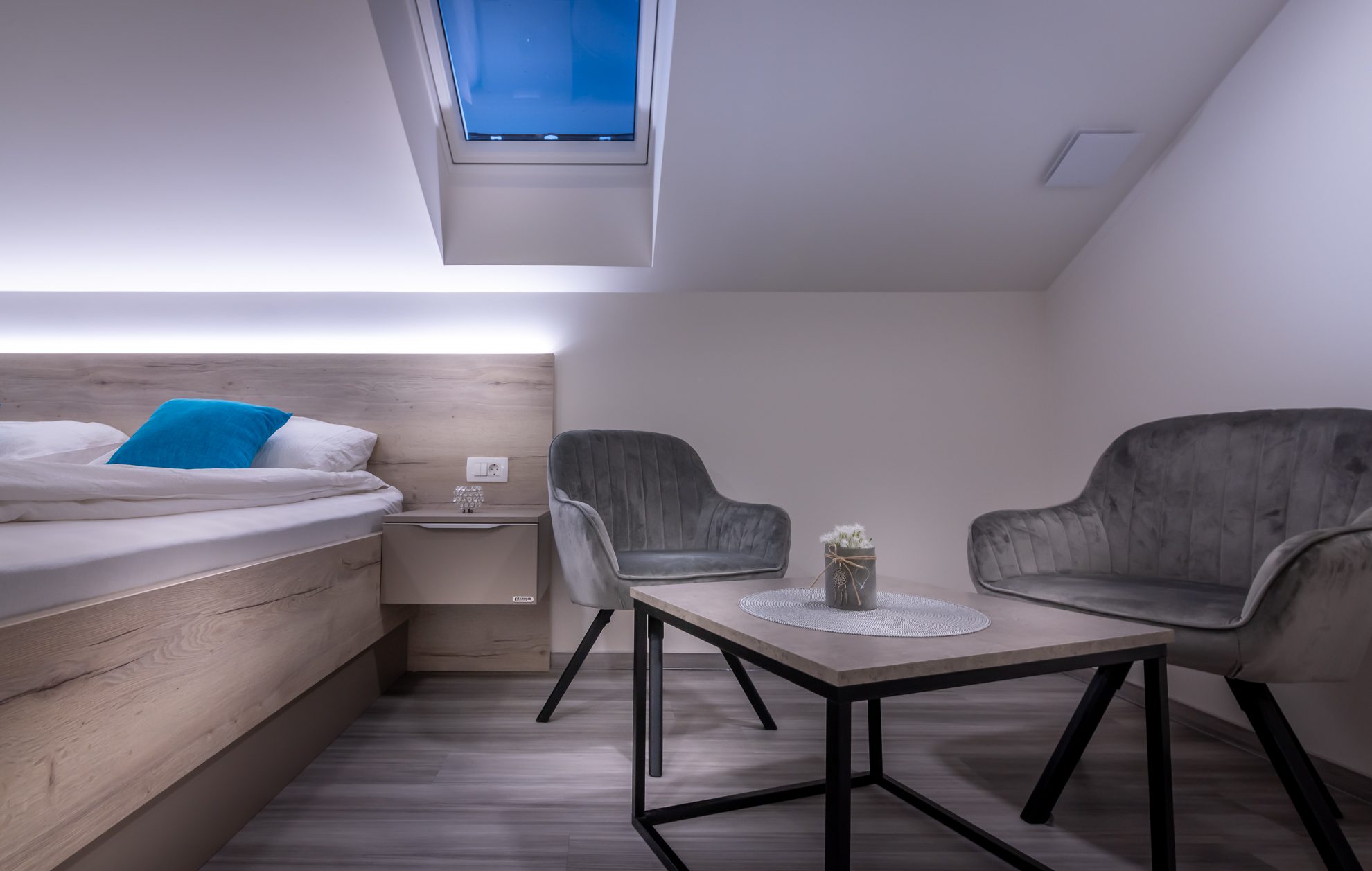 Udobna soba S-Center s posteljo, kotičkom s fotelji in strešnim oknom v mehki svetlobi